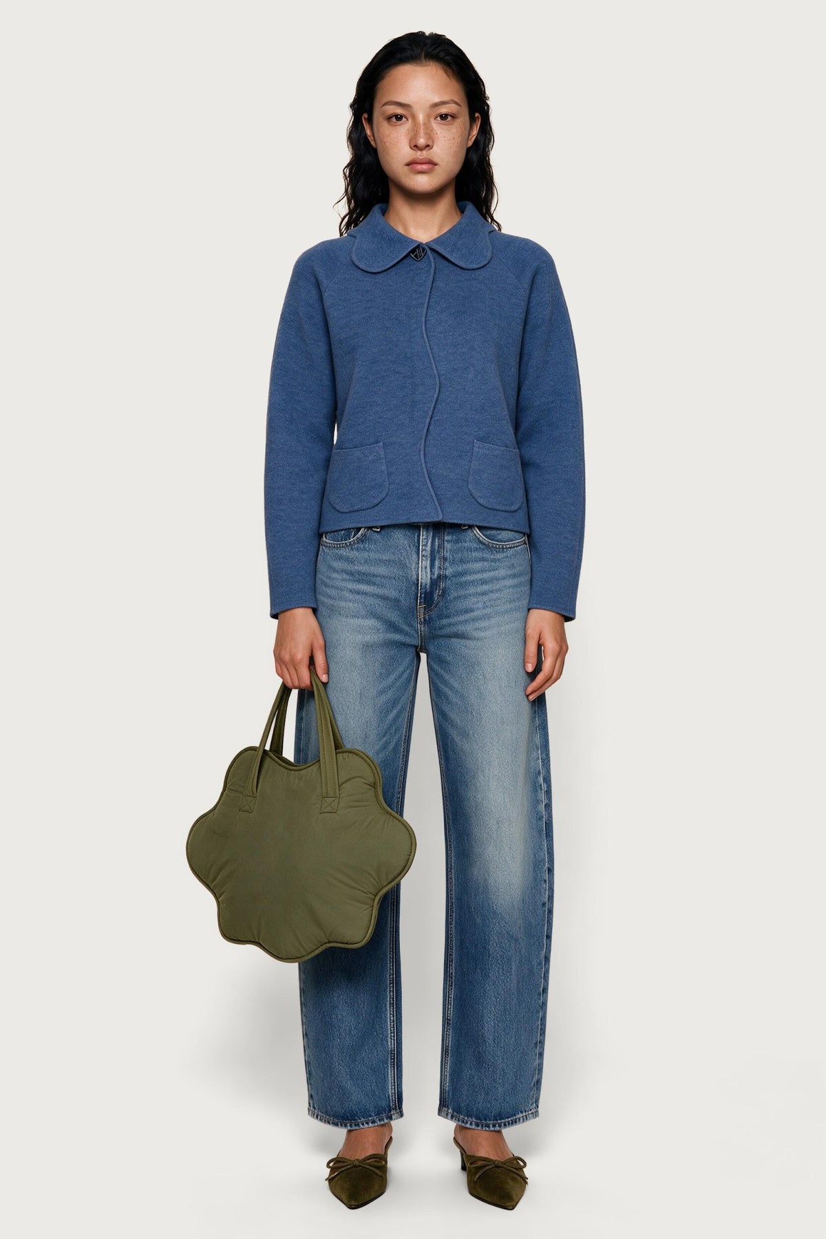 A model stands against a light gray studio background wearing a blue knit collared cardigan, blue jeans, olive green shoes, and holding an olive green cloud-shaped handbag.