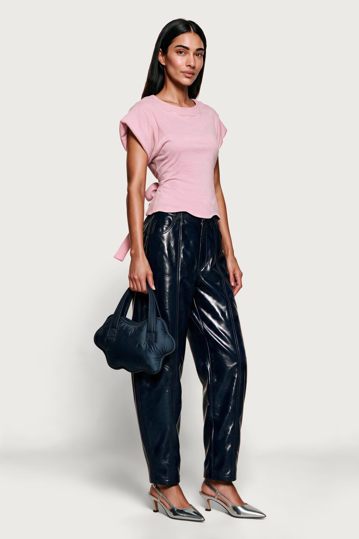 A model stands against a light gray studio background wearing a light pink short-sleeve top, shiny dark navy pants, silver pointed heels, and carrying a dark navy handbag.